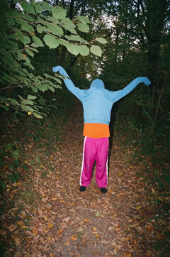 A flash-lit film photo of a figure with a sky blue hoodie pulled over their head, arms out, on a leafy forest path.