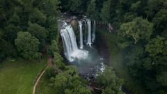 An aerial view of a waterfall cascading through a dense, verdant forest into a misty pool.